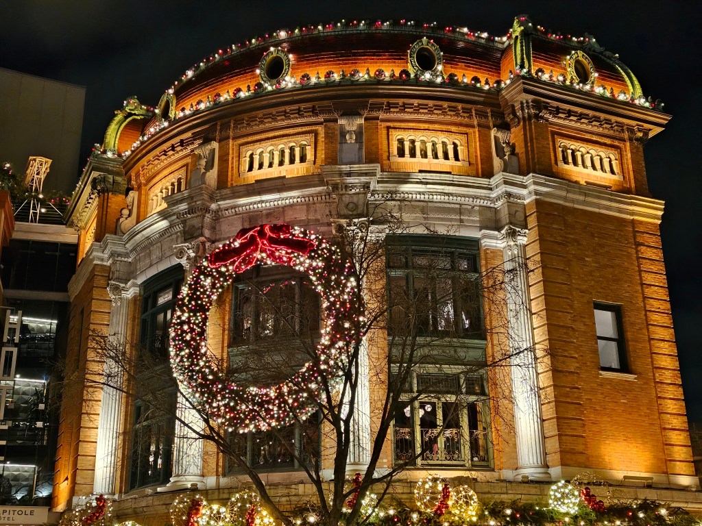 Large wreath made of lights attached to a building lit up at night with Christmas lights.